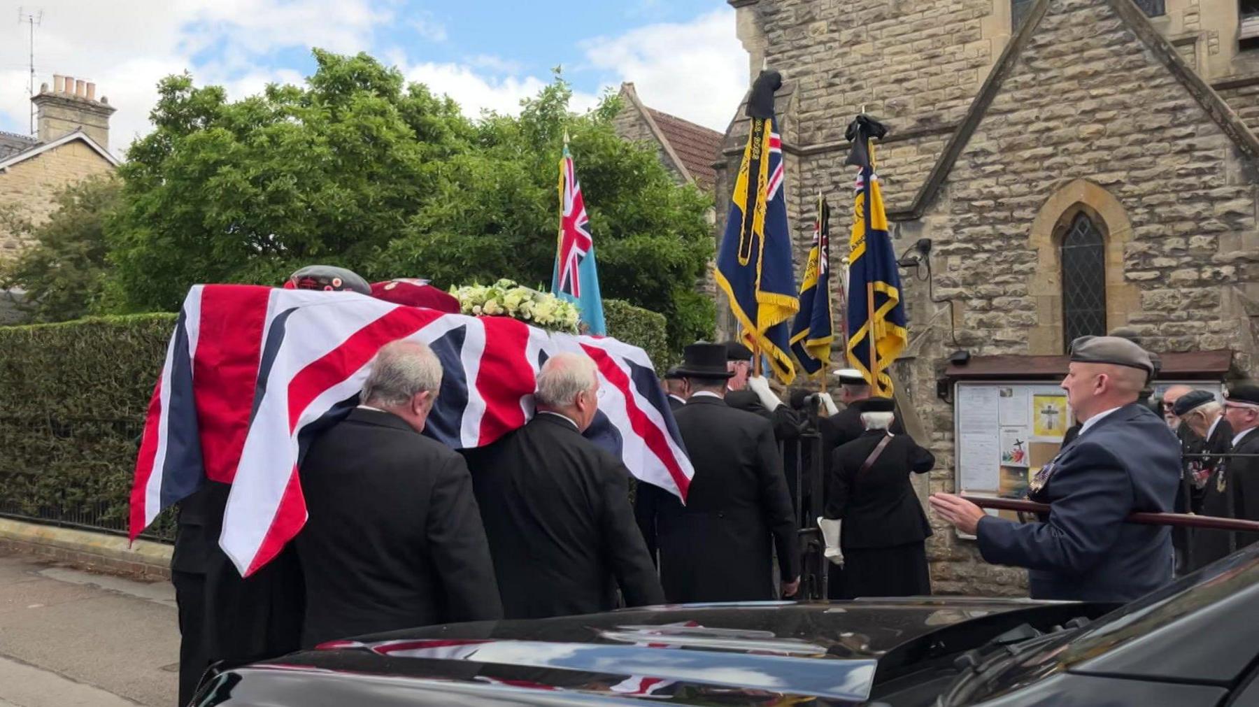 A coffin, draped in the Union Flag, is carried into a church by pallbearers wearing black suits as flags are waved in front by cadets.
