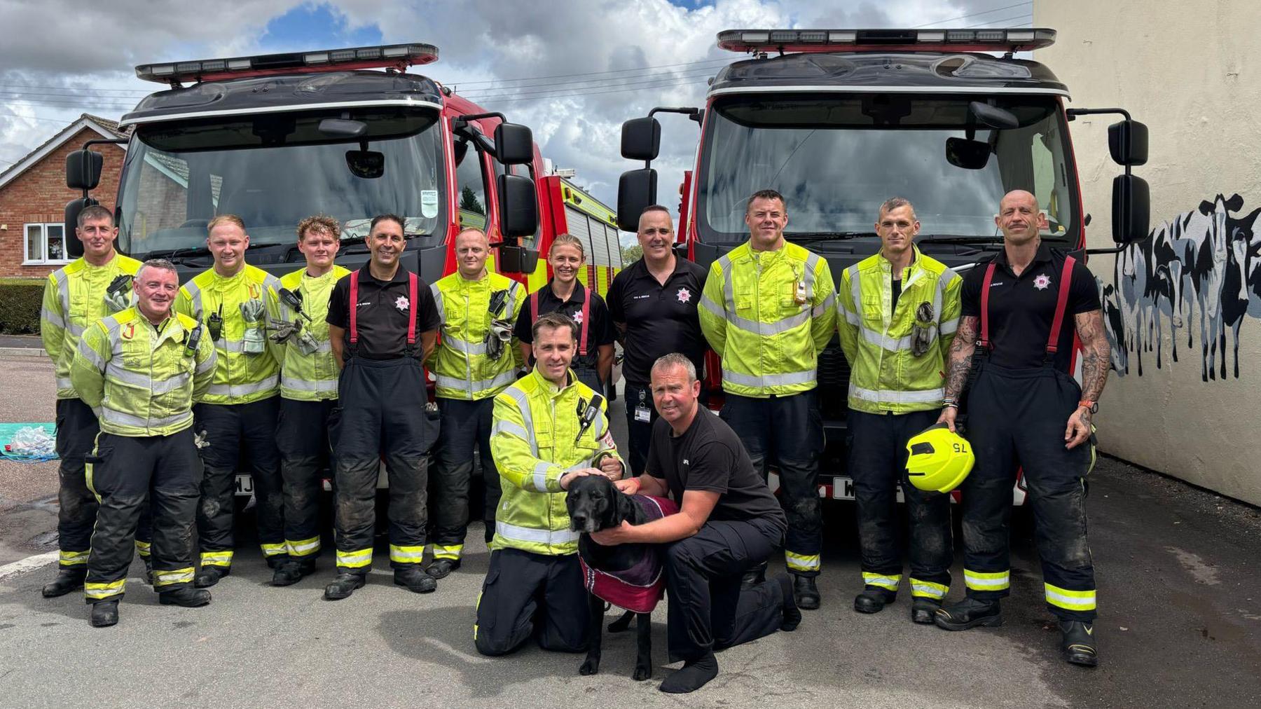 A group photo of firefighters in their uniforms. In the front of the group is a black labrador. They are all standing in front of two fire engines.