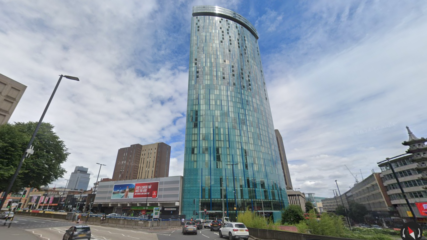 A large skyscraper covered in windows on a busy city street. Other skyscrapers can be seen in the background and cars are on the road below.