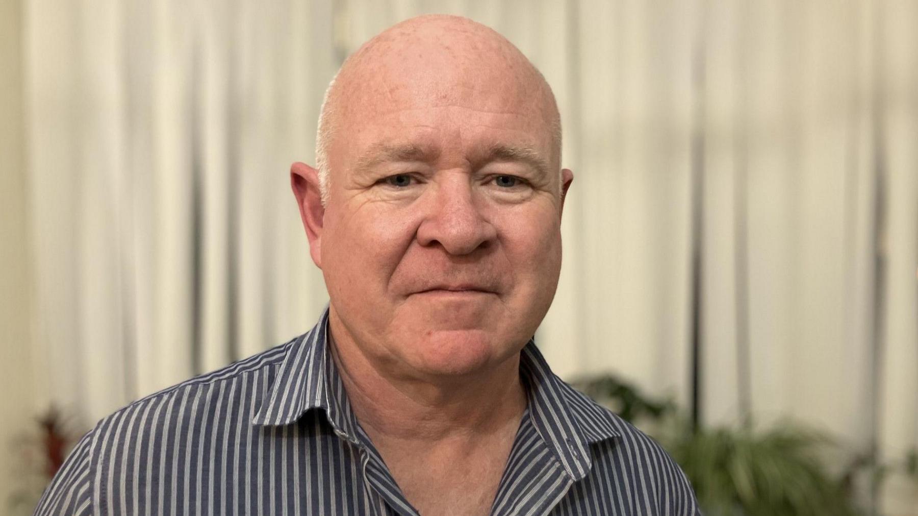 A head-and-shoulders image showing Steve Foster looking directly at the camera. He is semi-bald, with closely cropped white hair. He wears a striped blue and white shirt. He is standing in front of a beige window blind and there are pot-plants on a shelf behind him.