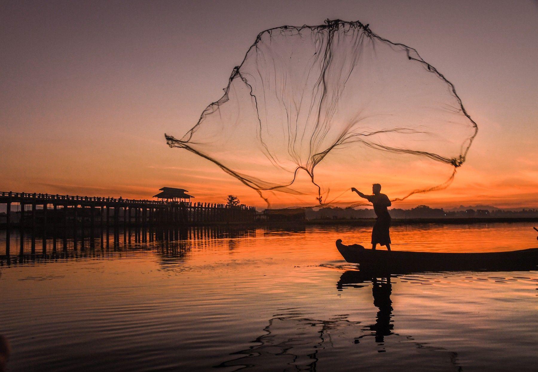 Silhouette of a man casting a large fishing net in the air from a boat during sunrise