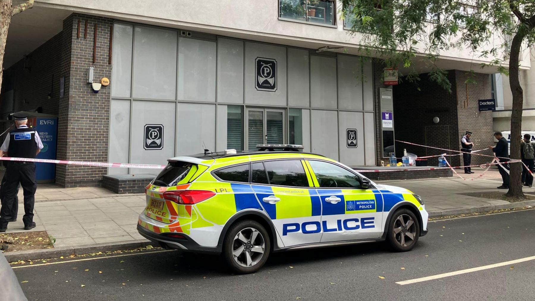 A police car parked outside a grey building and several police officers nearby behind police tape