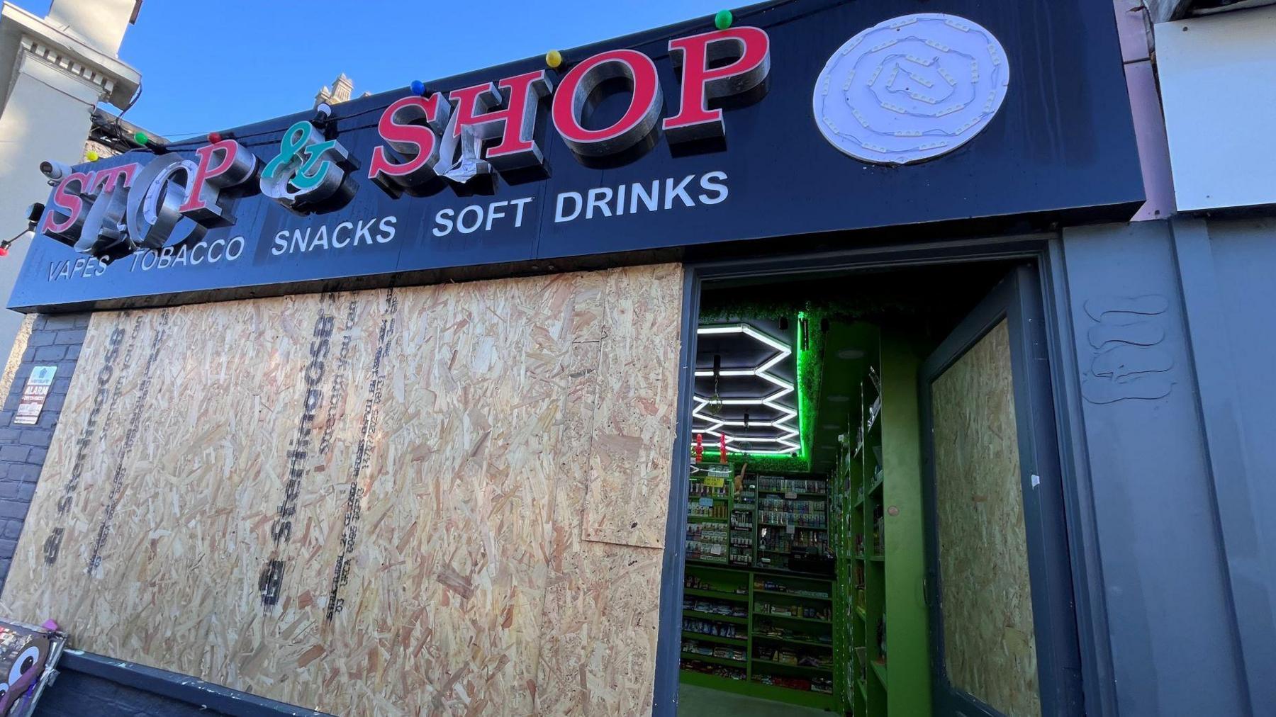 Shop windows boarded up. The entrance is open and neon lights are on inside. Vapes and cigarettes are on display at the back of the shop.
