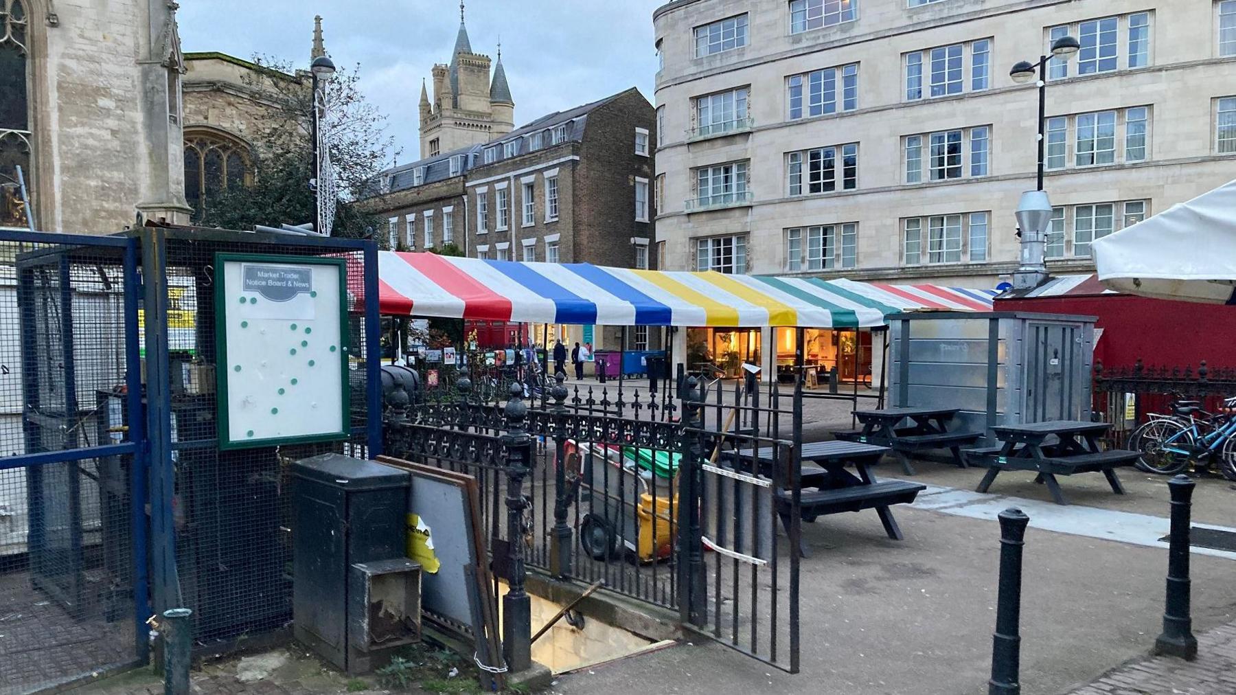 A scene in Cambridge city centre outside St Marys Church, Market Hill. There are no people around. There are coloured stripey awnings over a long market stall, some bikes locked up, and larger historical commercial buildings on the right. In the foreground are wrought iron gates, open, leading to a staircase  below ground.