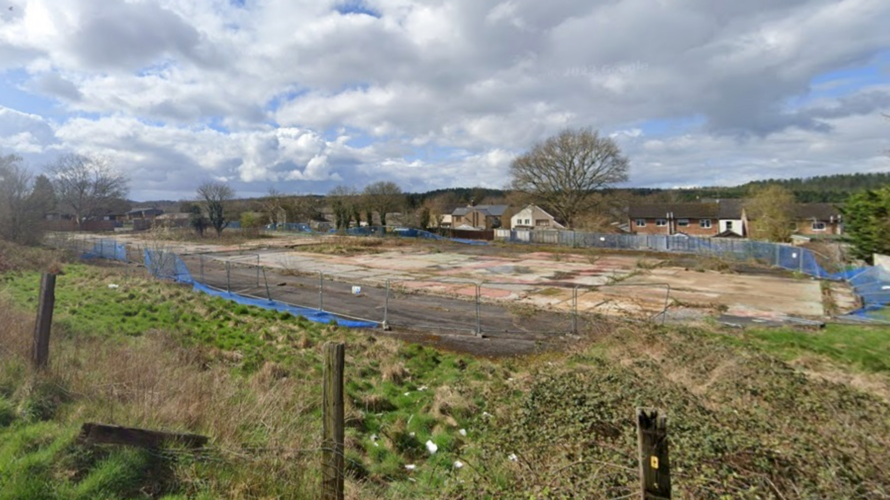 The GIS site in Cinderford. There is grass bordering the concrete site which is blocked off by grey fencing and blue netting. There are brown houses behind the site.