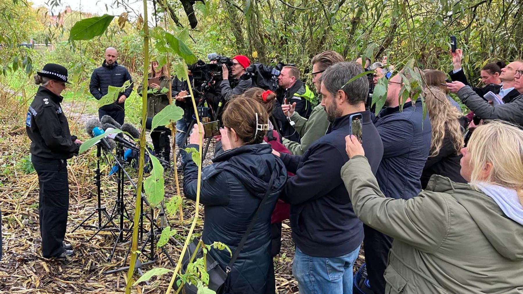 A group of people, some holding mobile phones, listening to a police officer speaking in front of some microphones