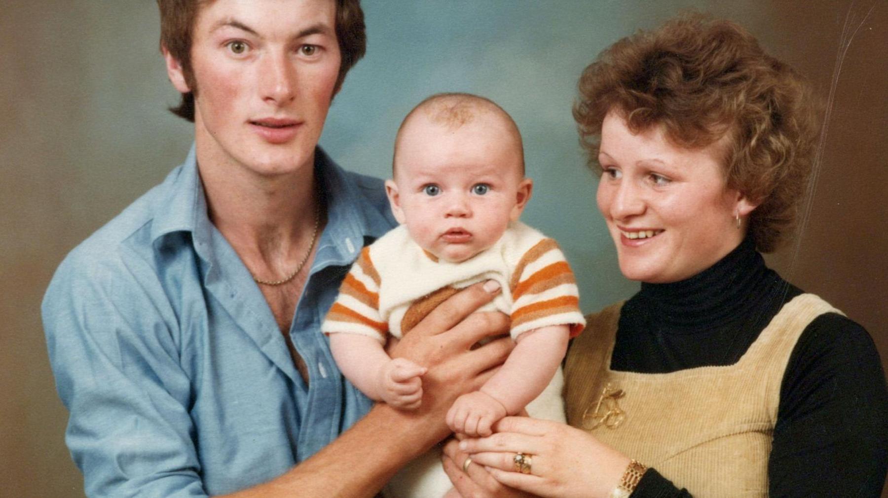 Luke Evans as a baby being held by his parents David and Yvonne. David is wearing a blue shirt, Yvonne is wearing a black polo-neck and a sandy coloured pair of dungarees. Luke in the middle is wearing a mustard and off-white babygro. 