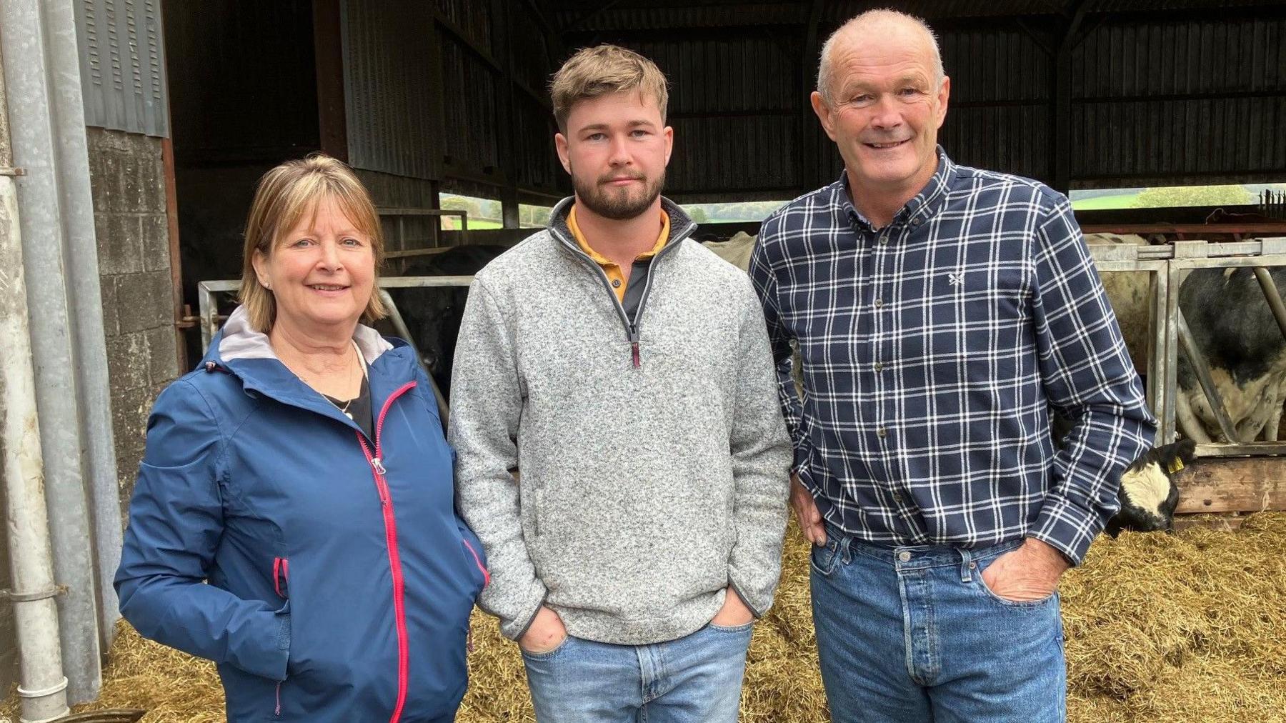Llun o'r teulu Evans yn sefyll o flaen ei ysgubor gwartheg. Daniel y mab sydd yn y canol yn gwisgo siwmper lwyd a jîns glas. Mae ganddo wallt brown a barf brown hefyd. I'r chwith mae ei mam, sy'n gwisgo cot law las. Mae ganddi wallt melyn. I'r dde mae ei dad Dai, sy'n gwisgo crys siec glas tywyll a gwyn a jîns glas.