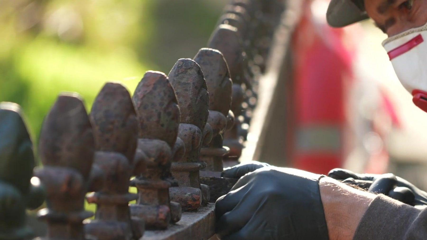 A line of small egg-shaped structures atop the railings of a bridge. A gloved and masked man is doing some work to the railings.