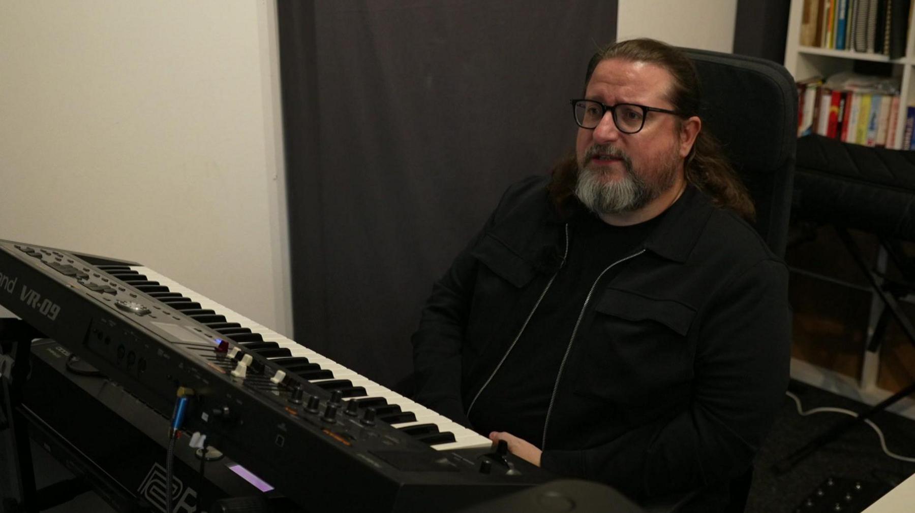 Barry Dallan, who is bearded, has a ponytail and wears glasses, is sitting in front of a keyboard in his rehearsal room in Liverpool