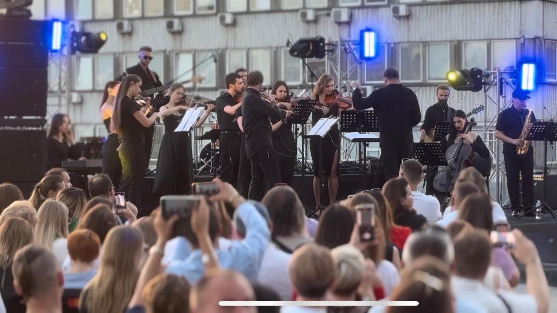 An orchestra plays on a rooftop as a crowd watches on, several people holding up phones to take photos and videos.