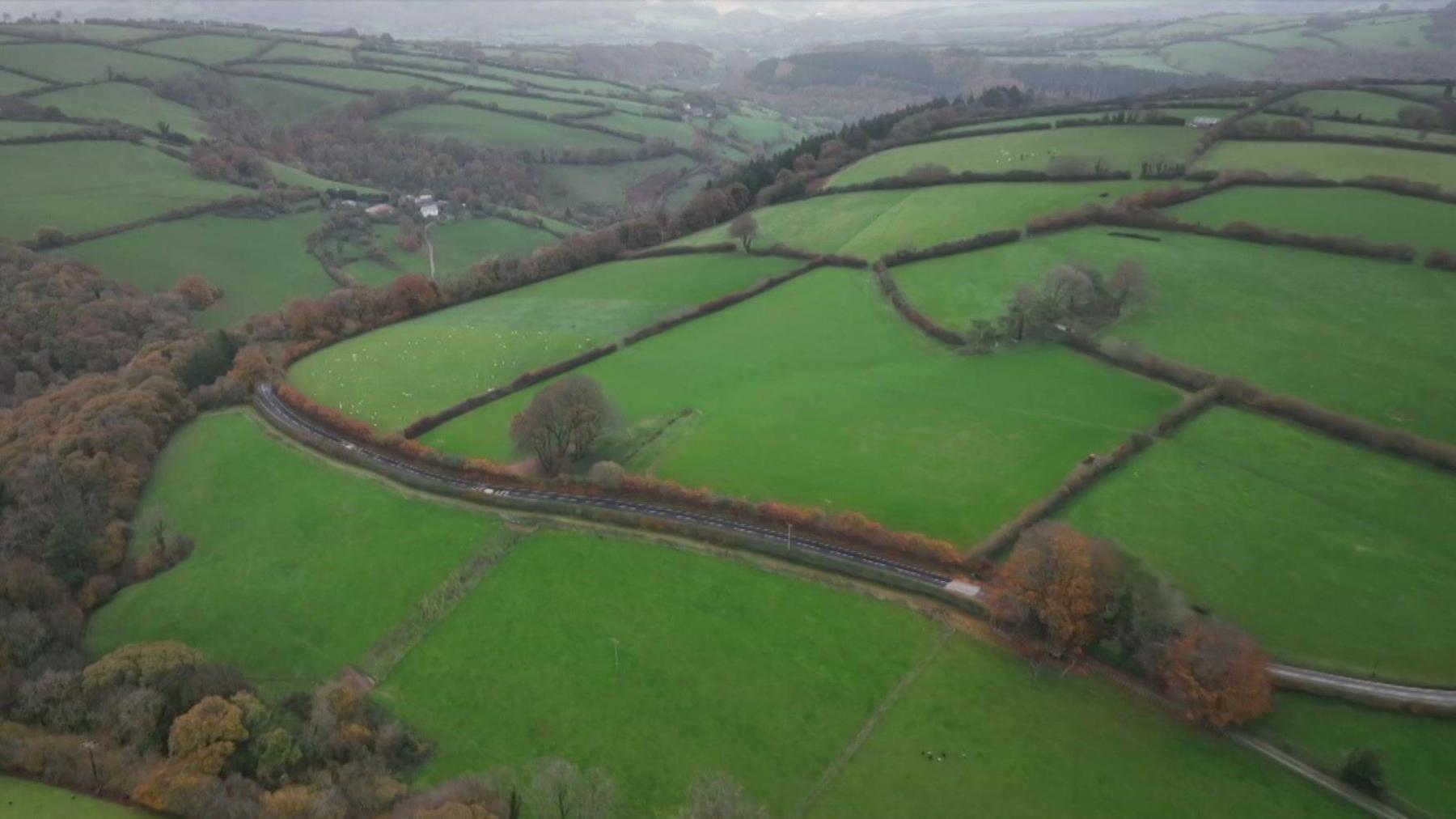 A drone shot showing a police roadblock on the A396 near Wheddon Cross in Somerset. The area is rural and the road is winding. There are many fields and trees across the landscape.