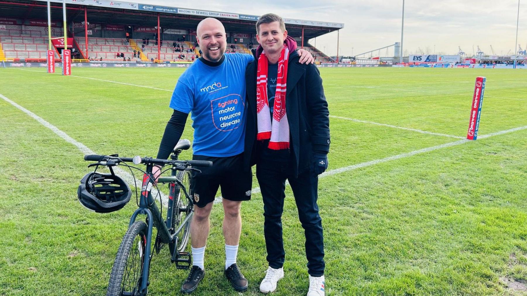 Liam Foster standing by his bike on a rugby pitch wearing a blue MND fundraising top, with Craig Eskrett wearing a red and white Hull KR scarf