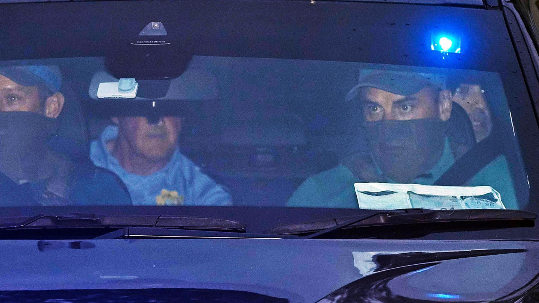 A close up image looking through the front windscreen of a vehicle. The two men in the front passenger seats are wearing baseball caps and have the bottom half of their face covered. There is a man on the left rear passenger seat wearing a blue shirt with a Gardai emblem. His face is obscured by the rear view mirror. On the right the face of Daniel Kinahan can be seen over the shoulder of one of the front passengers. He has stubble and is looking out the window.