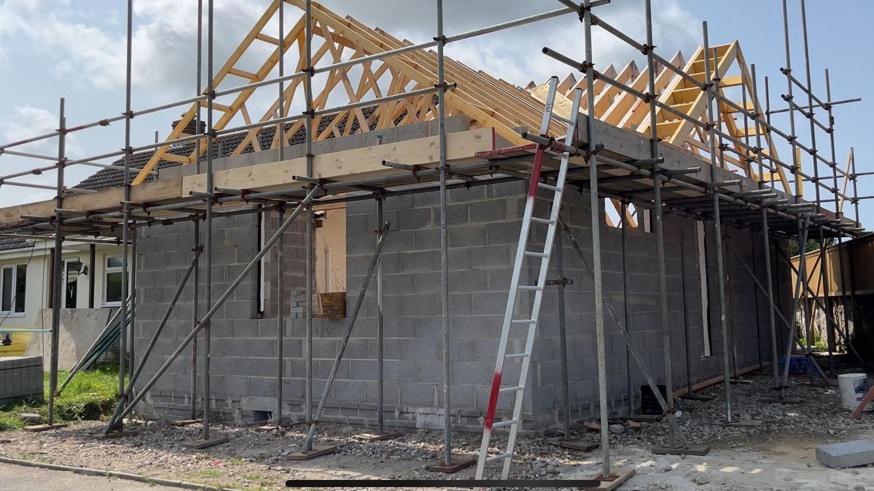 A brick work house with a timber roof, surrounded by scaffolding poles and with a ladder leading to the top of the scaffolding. 