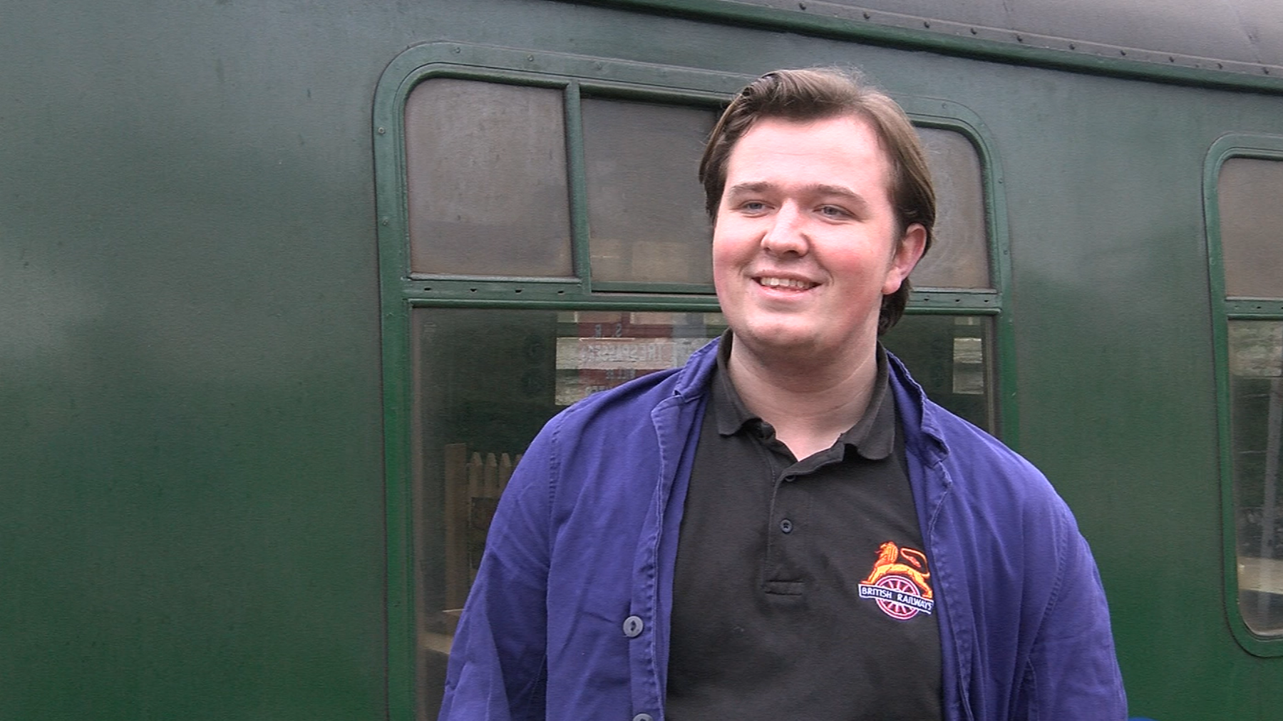 A young man wearing a black polo shirt and a blue overshirt standing in front of a green train carriage.