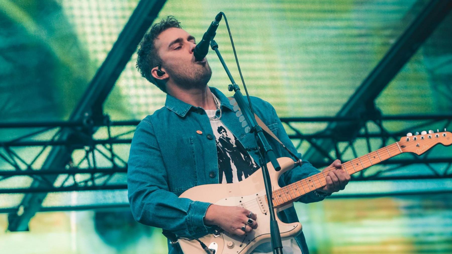 Sam Fender performing on stage at St James' Park. He has short dark hair and a dark stubbly beard, and is wearing a blue denim jacket with a white t shirt sporting black figures on it. He sings into a microphone while holding a guitar. There are green and yellow lights on a screen behind him.