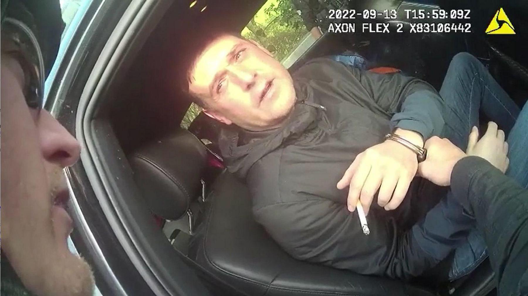 James Witham, holding a cigarette, sits in a car while a police officer places handcuffs around his wrists. He is wearing dark jeans and a hooded black jacket. 