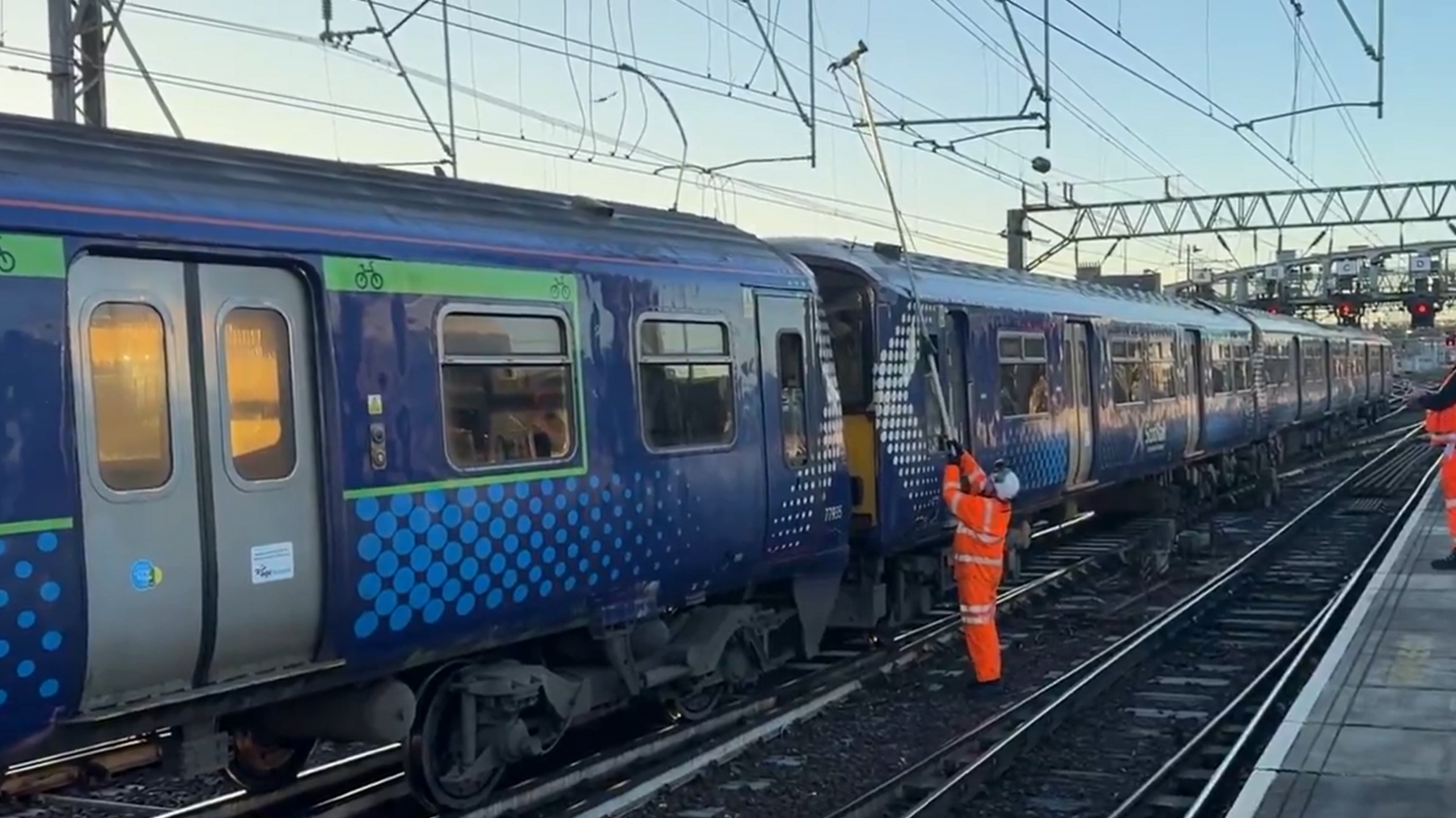 A railway worker in an orange overalls and a white helmet reaches above a blue train with a long pole to cut away at the damaged lines