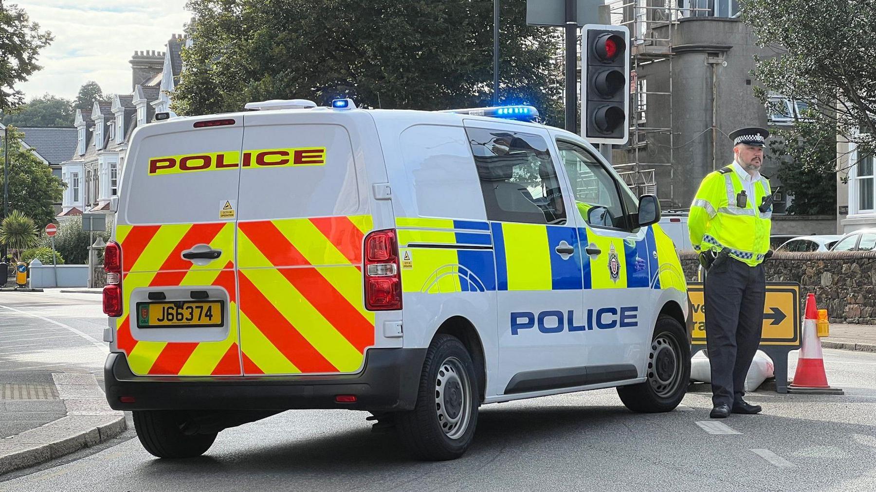 A police van is parked across a road junction and there is a uniformed police officer wearing a high-viz anorak standing with his hands behind his back near the vehicle. There is a traffic cone next to a road diversion sign. It is a residential area.