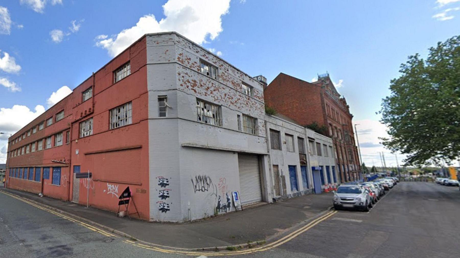 A large flat roofed warehouse building. It is painted orange on one side and grey on another. There is some graffiti on the walls