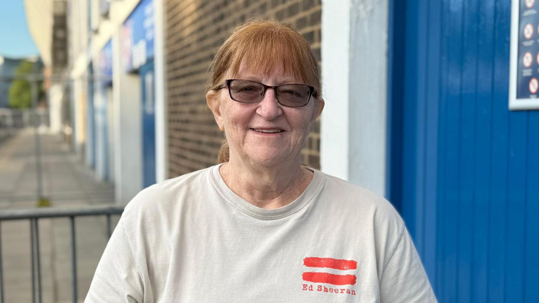 A head and shoulders image of Deborah Marshall He is wearing a white T-shirt and dark sunglasses. He is standing in front of Portman Road and looking into the camera.