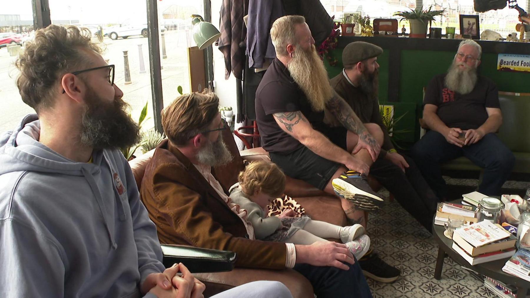 Five bearded men sat on chairs in a barber shop having a conversation.