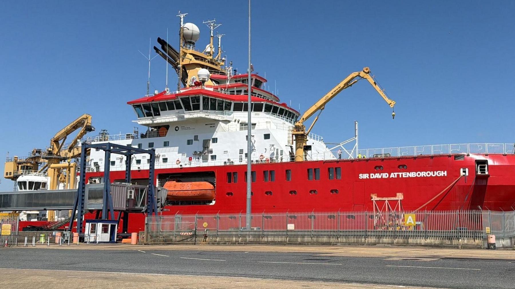 A ship is docked harbourside. It is mainly red-coloured with a white middle deck.