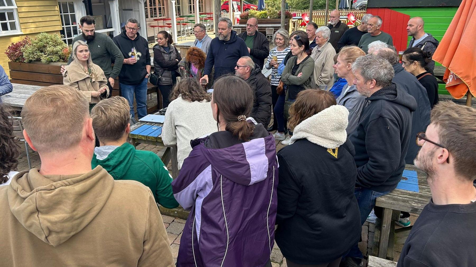 A group of people wearing coats and hoodies standing in a pub garden listening to a woman speak.