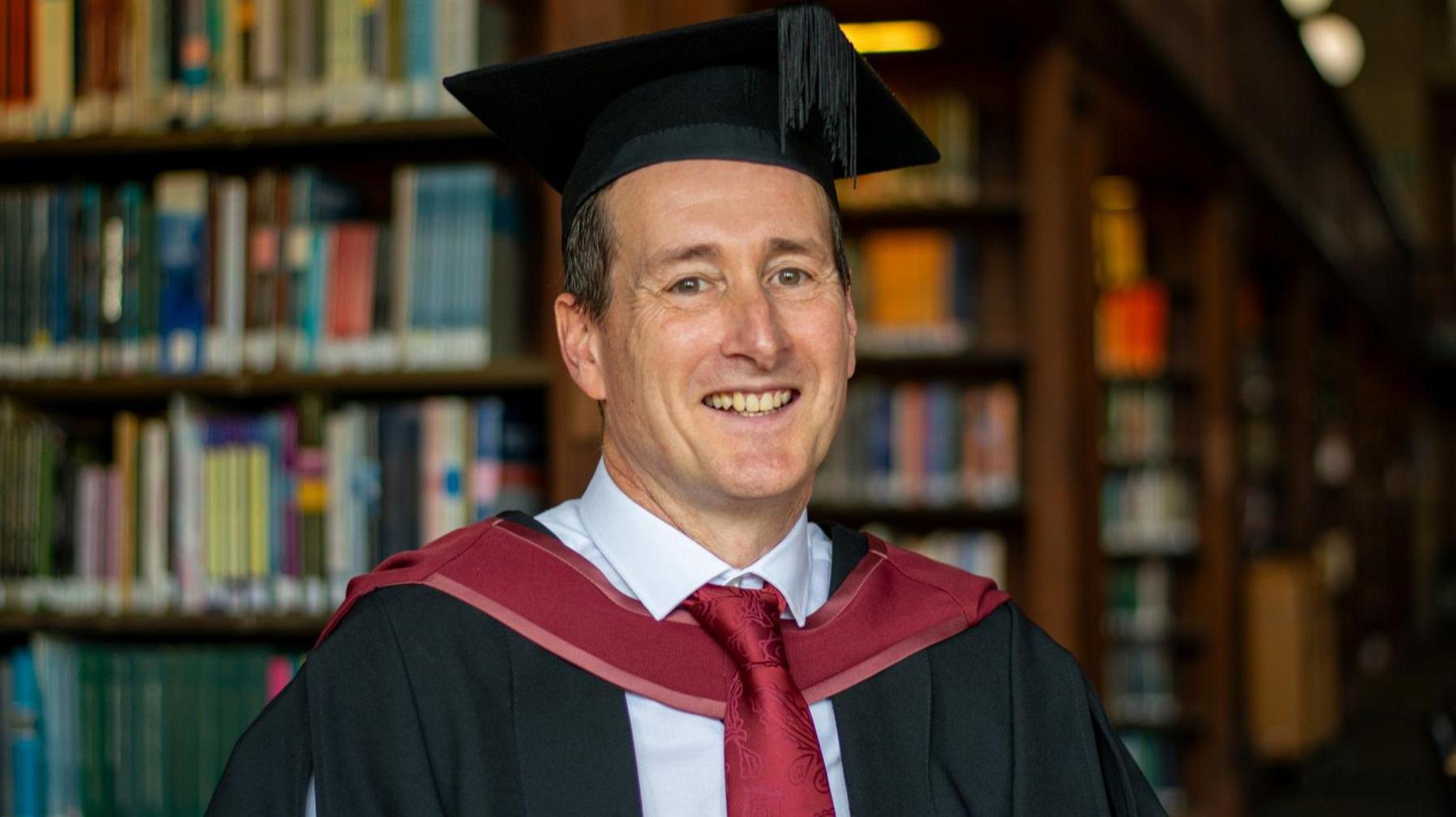 A man wearing black and red graduation robes pictured in front of a bookshelf