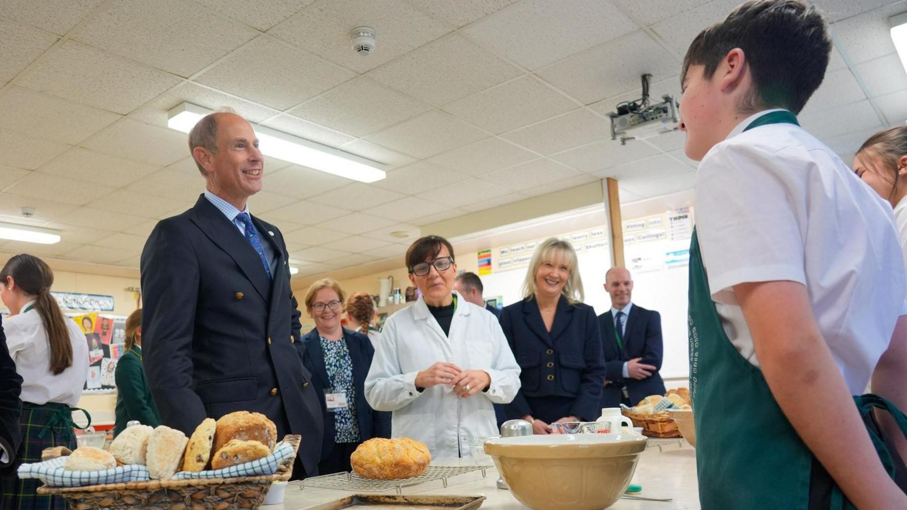 The Duke of Edinburgh, Prince Edward, standing at a school bench chatting to a pupil in a classroom, there are teachers and staff members standing around them. The pupil is wearing an apron and there is baskets of bread on the table and mixing bowls. 