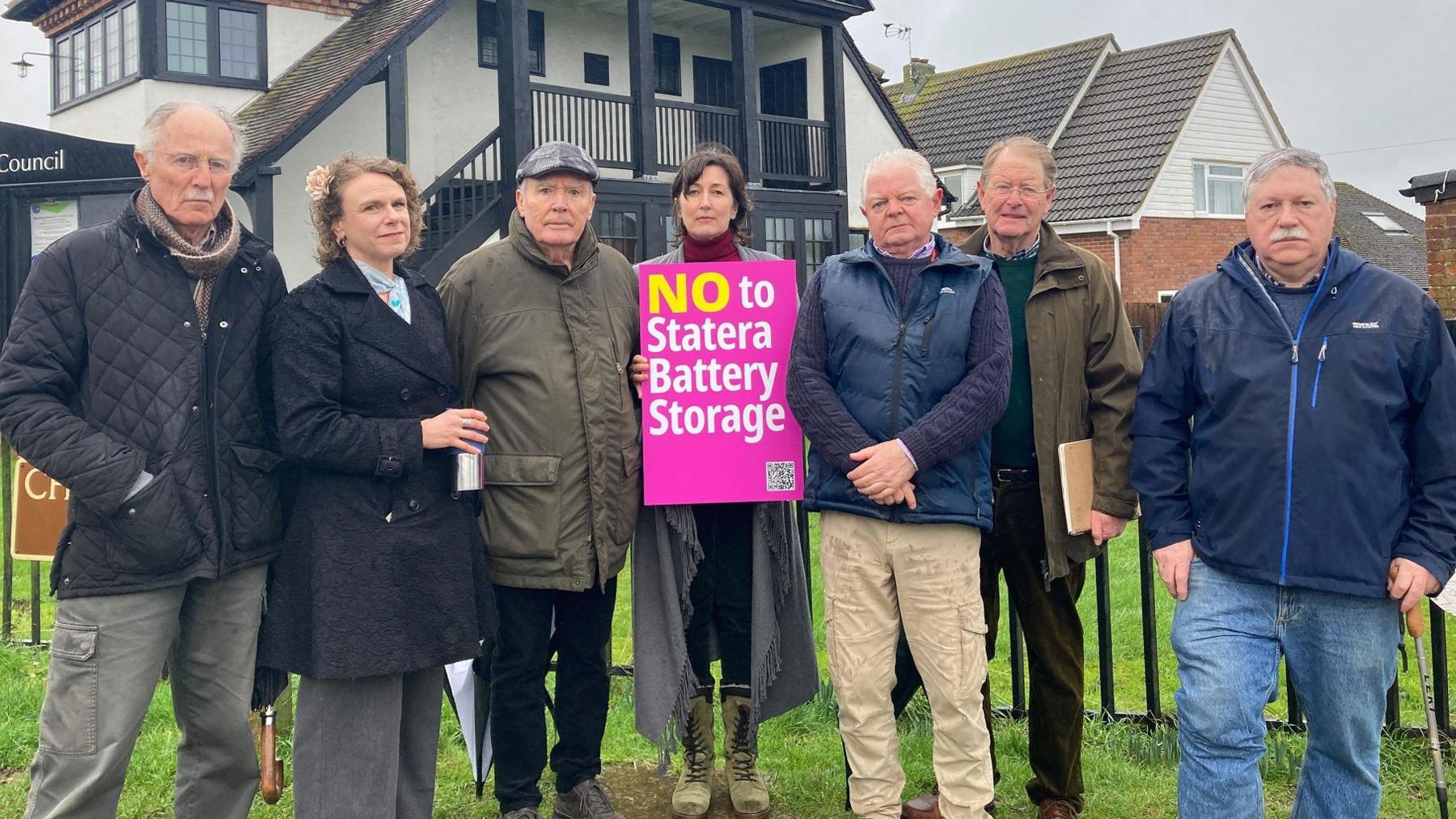 Seven people stand in a row outside a council office, in protest against the proposed battery site. A lady in the middle holds a bright pink sign saying "NO to Stater Battery Storage". There are two women and five men, all wearing winter coats.