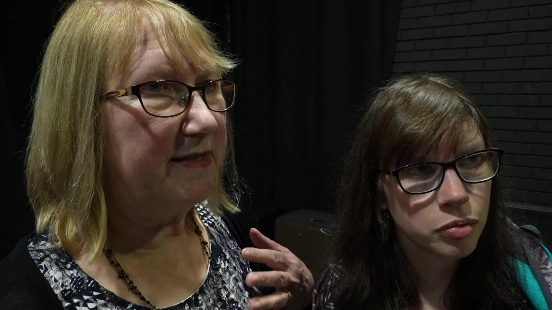 Hannah stands to the right and has long brown hair and wears glasses. Her mother, an older woman, stands to the left. She has blonde, shoulder-length hair and also wears glasses.