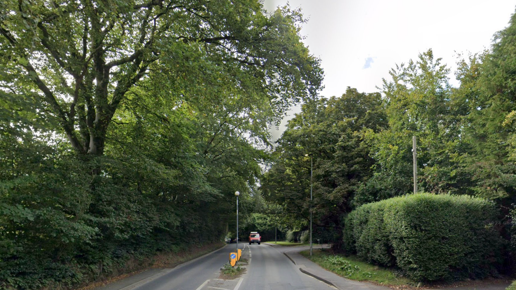 A road surrounded by trees with a red van coming on the right. Google maps image.