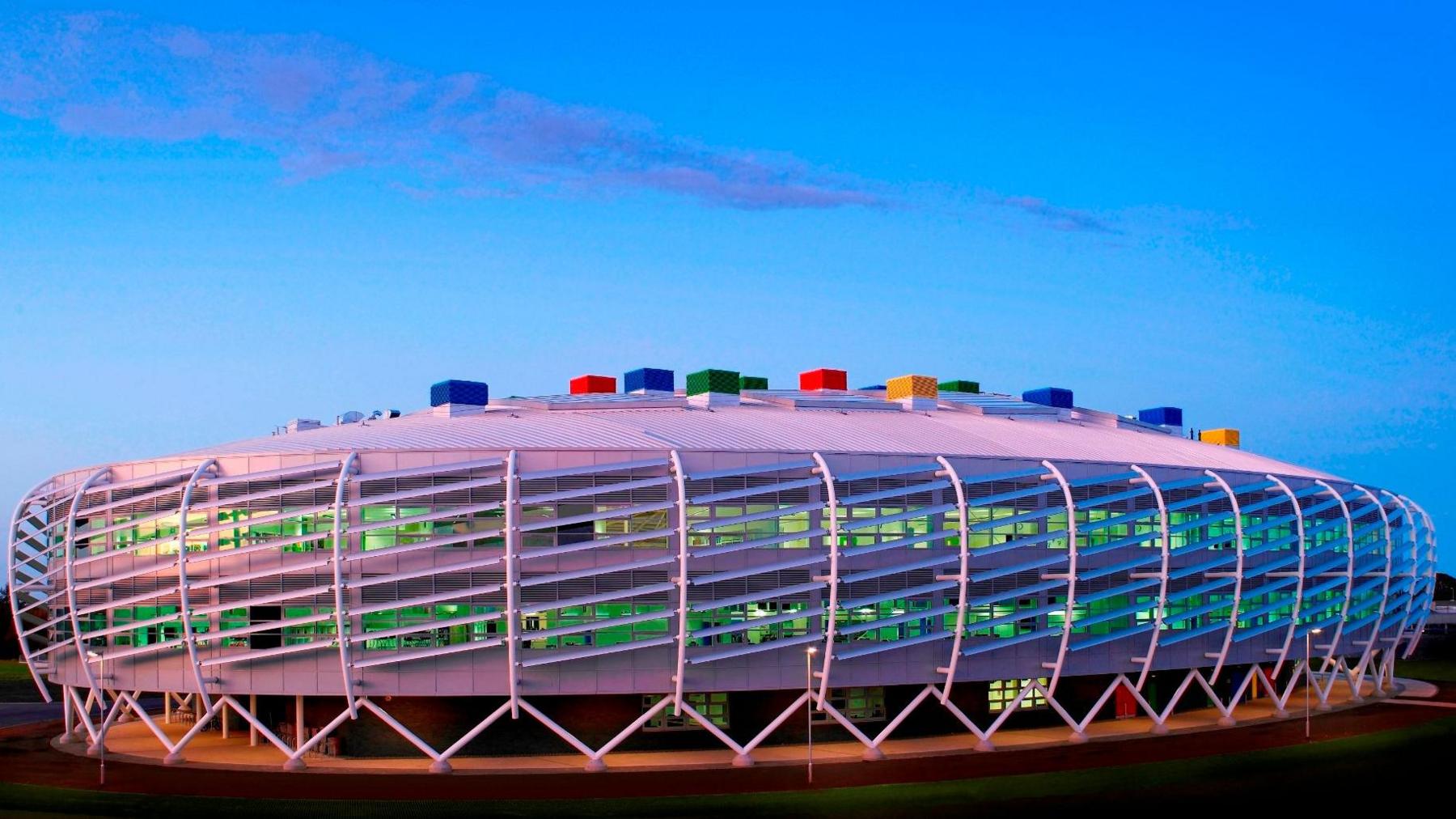 Colourful, oval-shaped, futuristic-looking Monkseaton High School which has multi-coloured blocks on its roof. The building is wrapped by white steelwork which sits on A-shaped white steel rods. Lights visible through windows make the rooms appear green coloured.