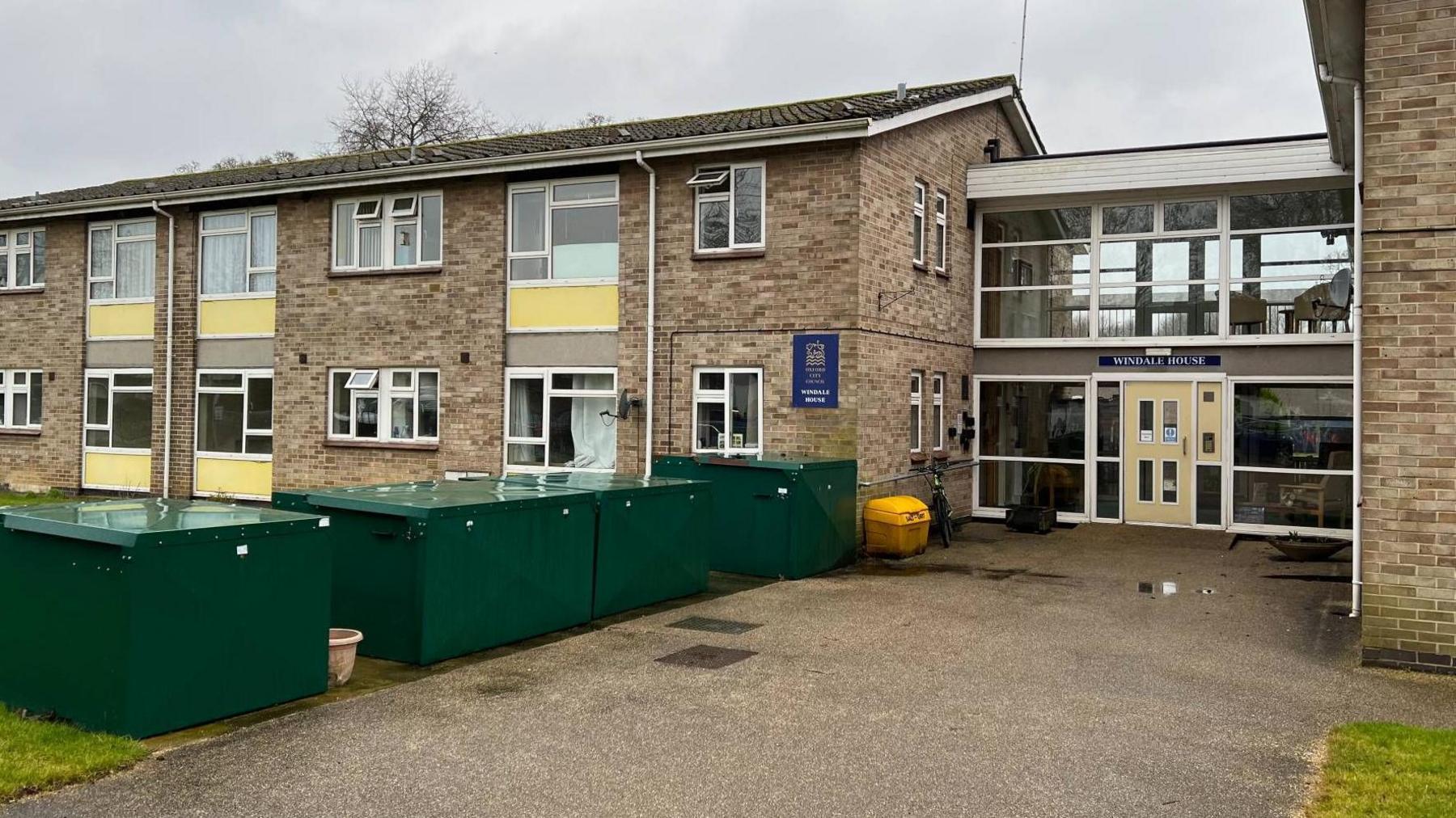 The entrance to Windale House, a two-storey brick building. Large green containers stand beside the entrance pathway, which leads to a doorway beneath a glass-fronted first-floor communal area with a table and chairs.