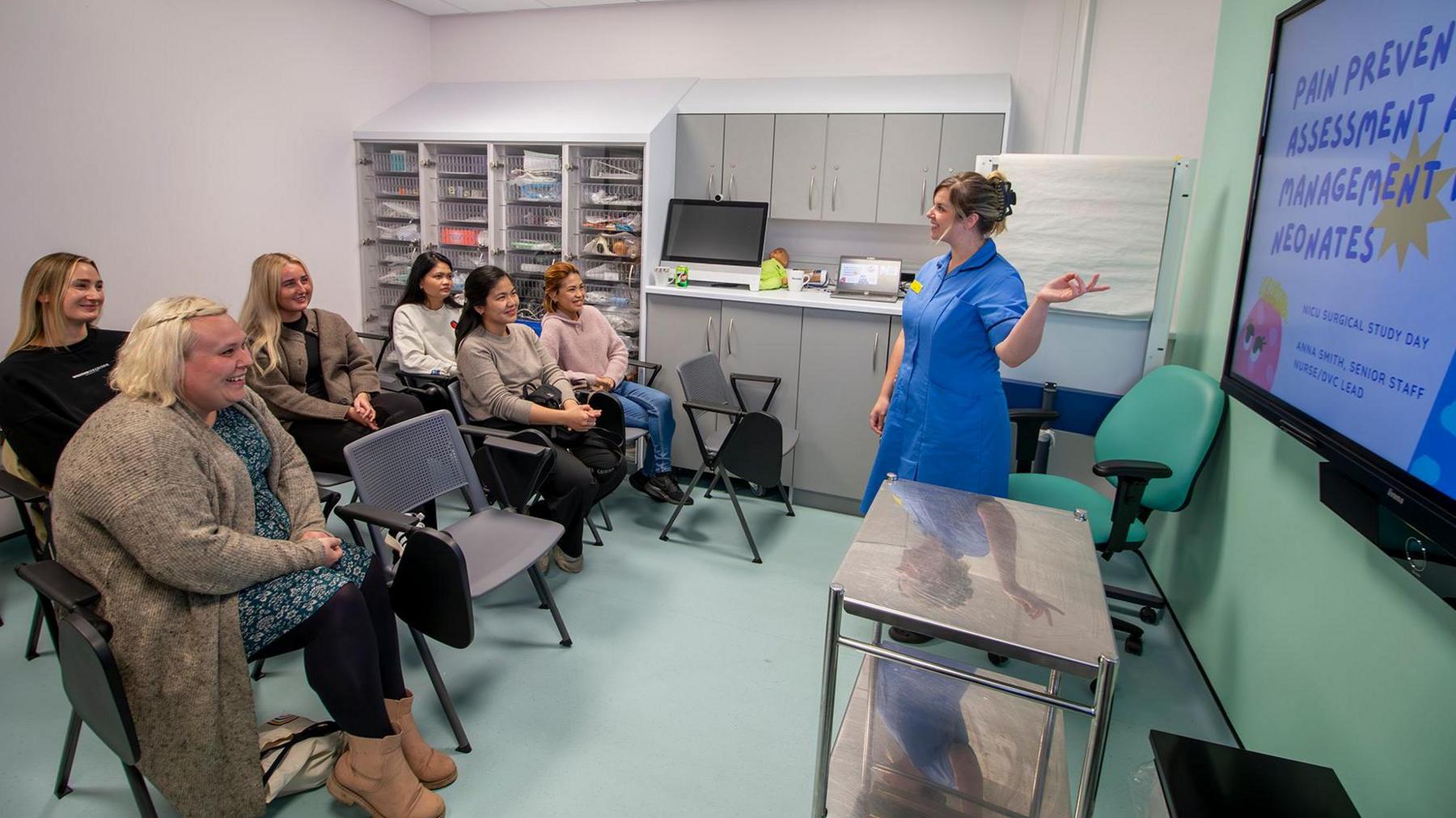 A midwife in a blue uniform is standing on the right smiling and pointing to a TV screen, which is displaying a presentation. Six women are sitting on two rows of chairs on the left smiling and facing the midwife and TV screen