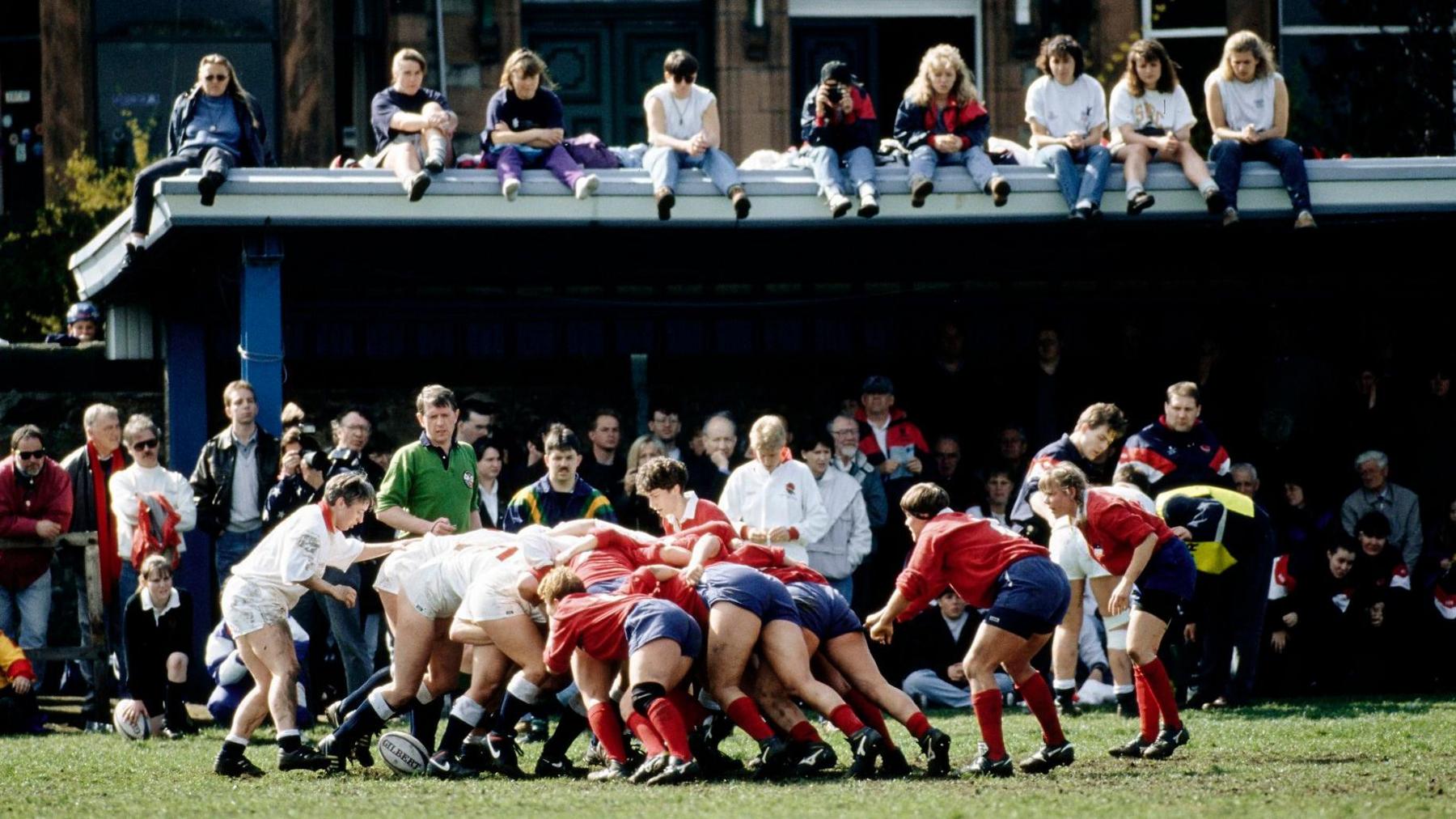 A scrum during women's rugby match in 1994, with players dressed in white taking on opponents in red and blue. They are watched by a crowd packed into a small stand, with some girls and young women watching while perched on the roof.