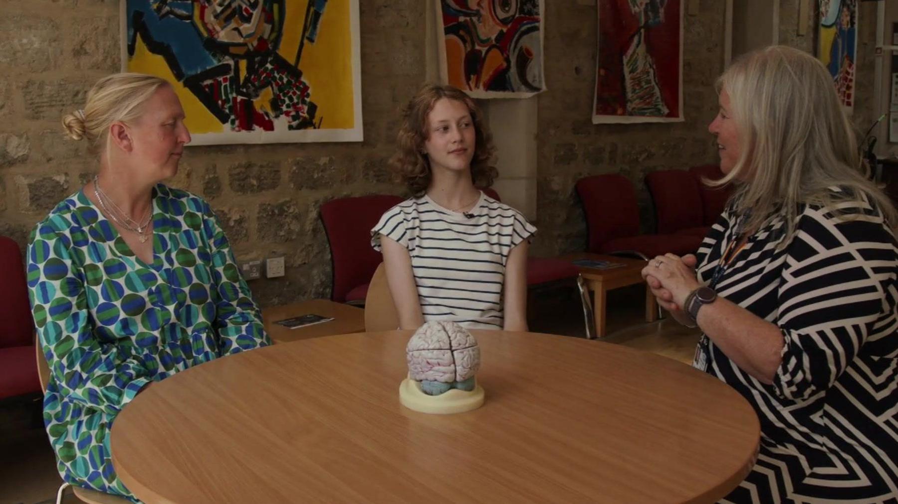 Josie, Bea and Prof Mackay sit round a round wooden table with a small cream sculpture at the centre. Josie is wearing a dress with a blue, green and white geometric circle pattern. She has blonde hair pulled back in a bun. Bea is wearing a blue and white horizontal striped T-shirt and Prof Mackay is wearing a dress with black and cream diagonal stripes