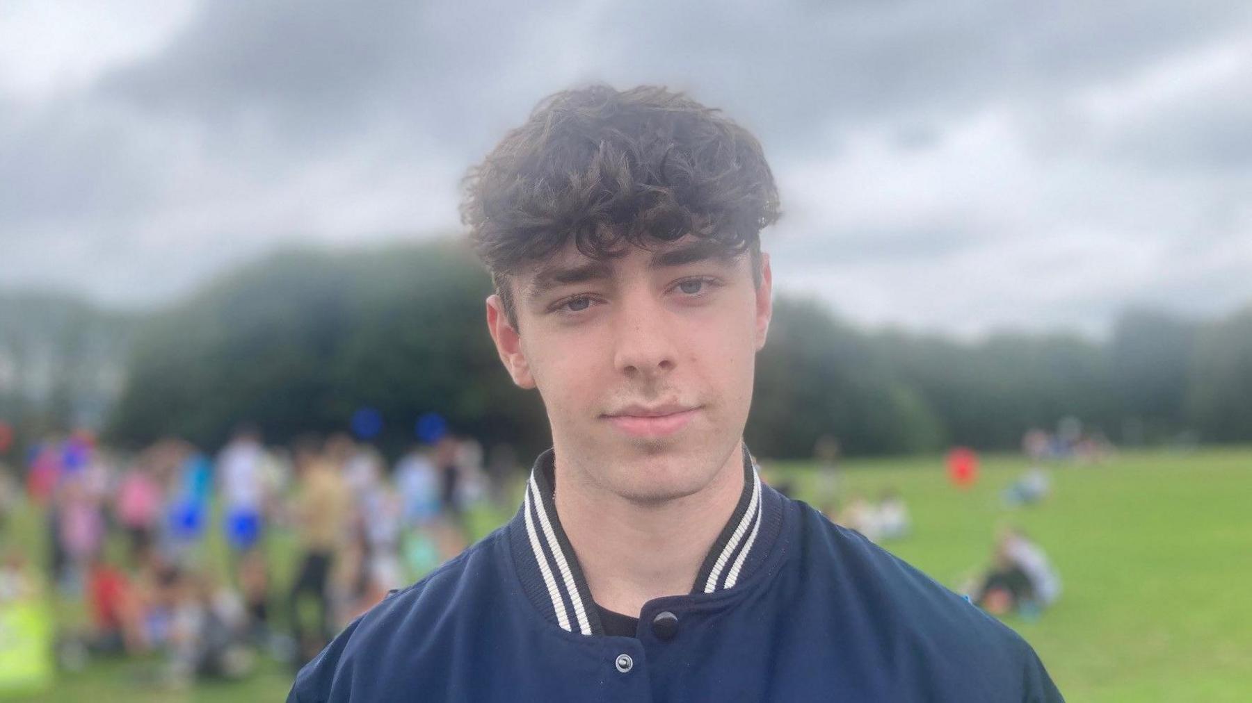 A man with curly brown hair looks at the camera. He is wearing a navy jacket. Behind him is a large field with a crowd of people.
