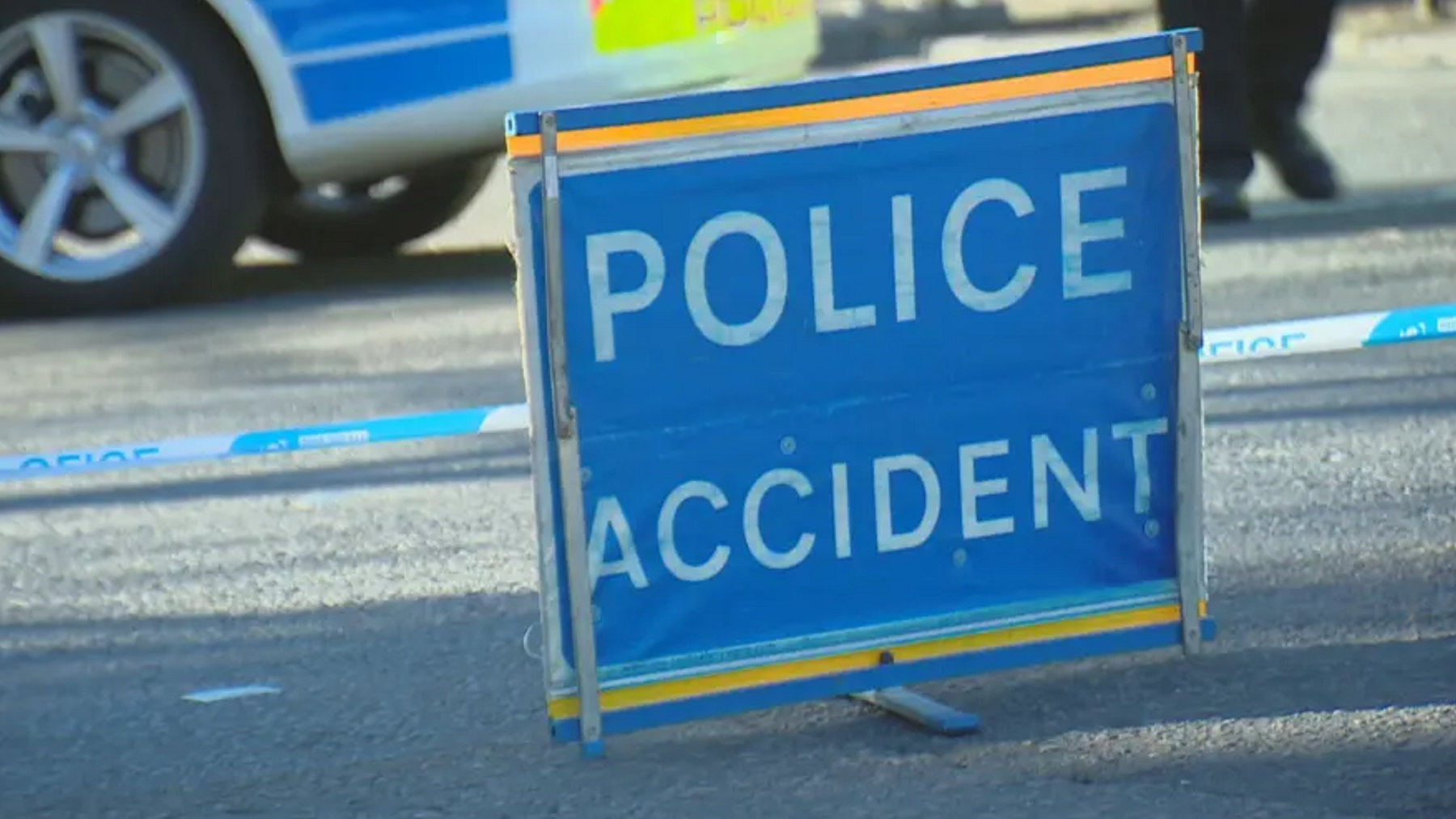 A stock image of a scene of a collision. A blue sign can be seen which reads police accident. Police tape also in the photo.