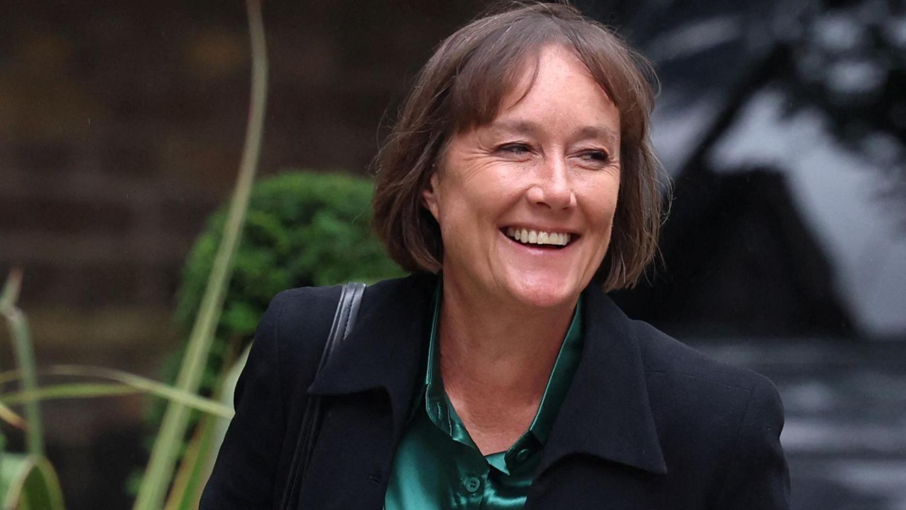 Jo Stevens in a black suit with a green satin shirt, she is smiling at the camera and holding a red folder