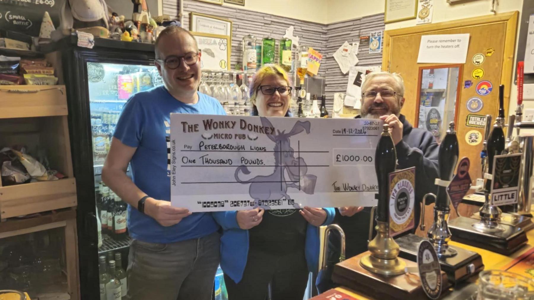 Two members of Peterborough Lions group on the left holding a big cheque of £1,000 with Dave Williams- standing behind a bar