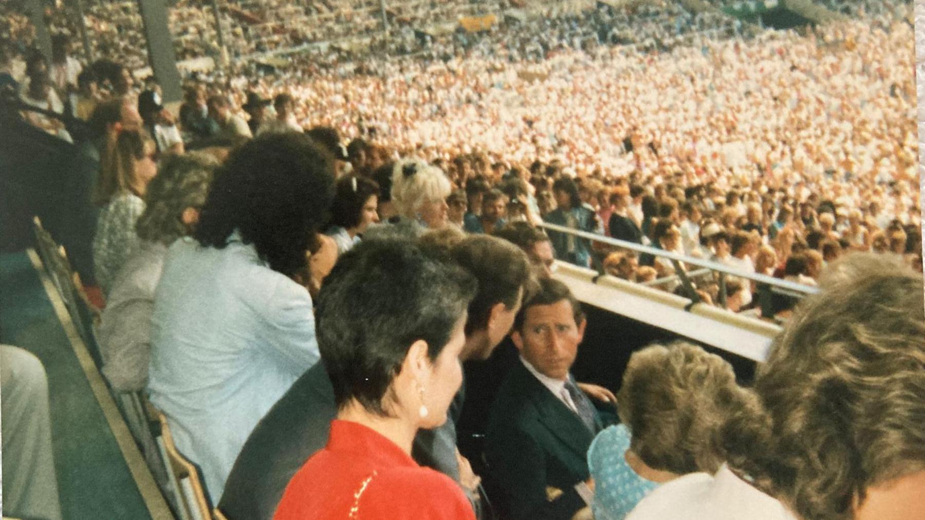 A picture of people sitting down in their rows watching on as a concert takes place, with a crowd in the background. One man, Prince Charles, can be seen turning his head and talking to someone behind him.