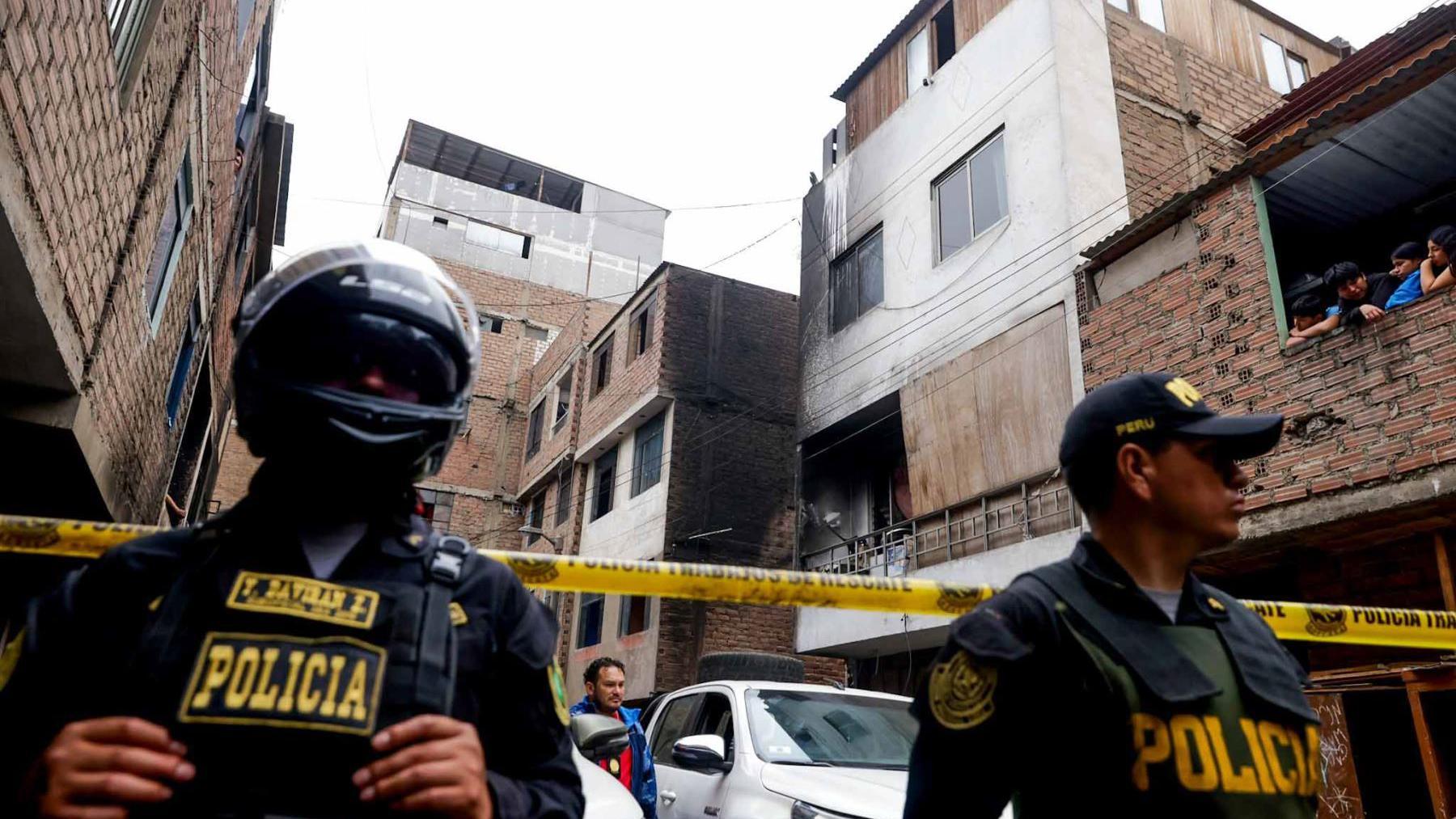 Two police officers guard a cordoned off area, which has several apartment blocks that are about four storeys high. One of them is blackened by fire.