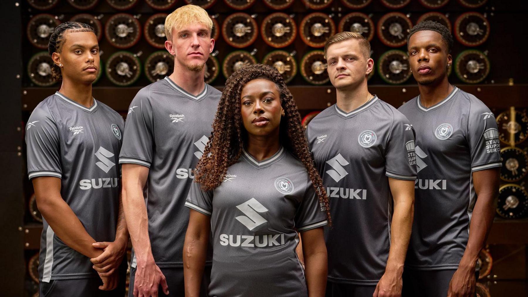 A group of people, of different eights and genders are stood in front of the Bombe machine wearing the new MK Dons football kits.