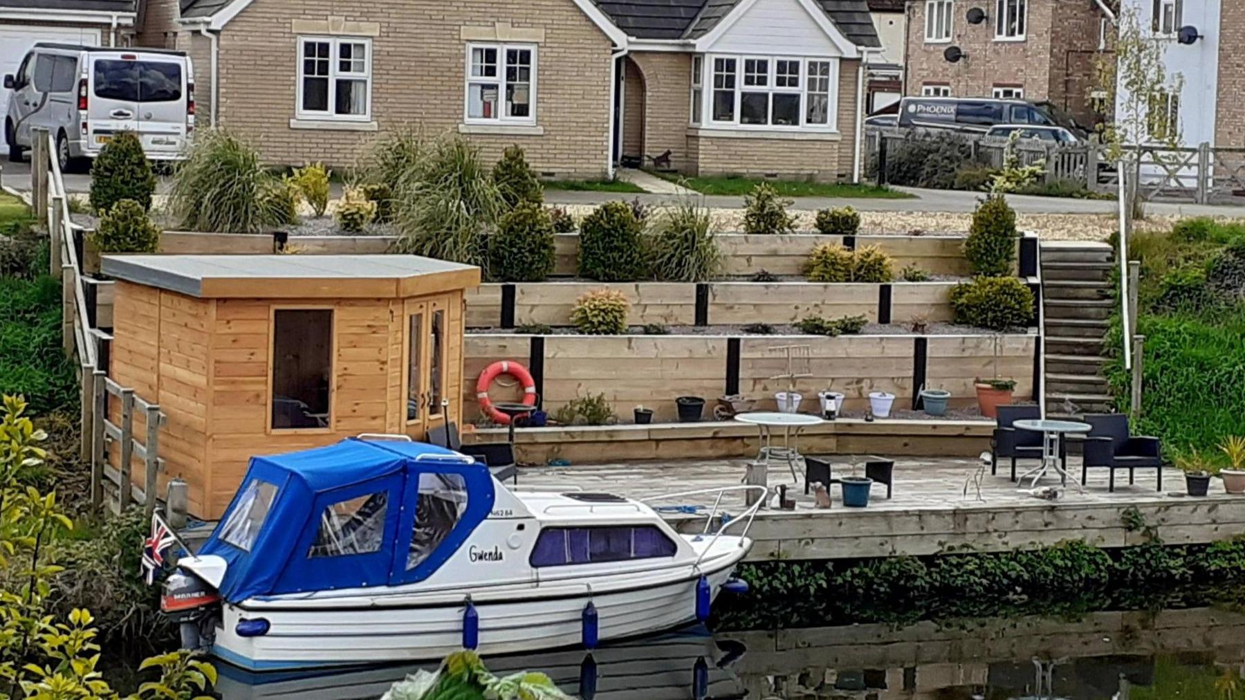 A 4.5m long (15ft) white cabin cruiser with a blue canopy. There is a grey and orange outboard motor at the back and the vessel is moored to a wooden decked area with a wooden summerhouse. Patio furniture, tables and chairs are on the right and there is a staggered raised bed effect leading up to the back garden of a modern bungalow with other homes around it.