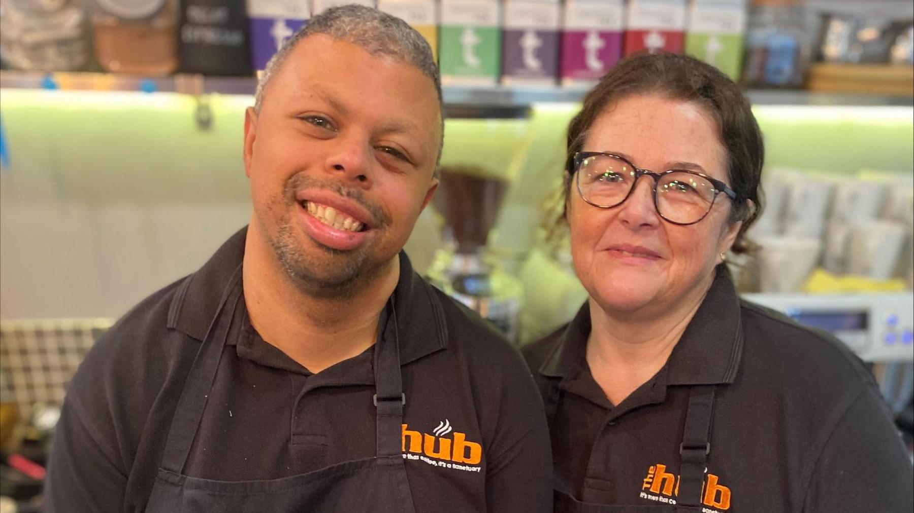 Reece and Penny standing next to each other, smiling. They are both wearing black uniforms saying The Hub in orange letters.