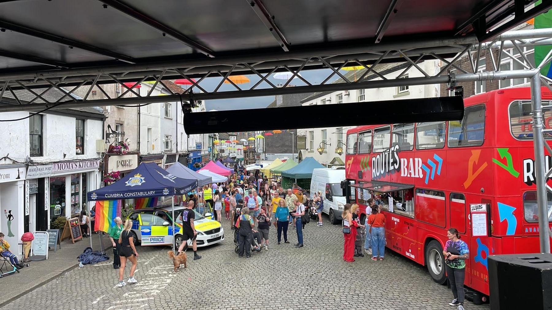 The photo, taken from a stage, looks out onto multiple brightly coloured stalls. There look to be about 100 people.