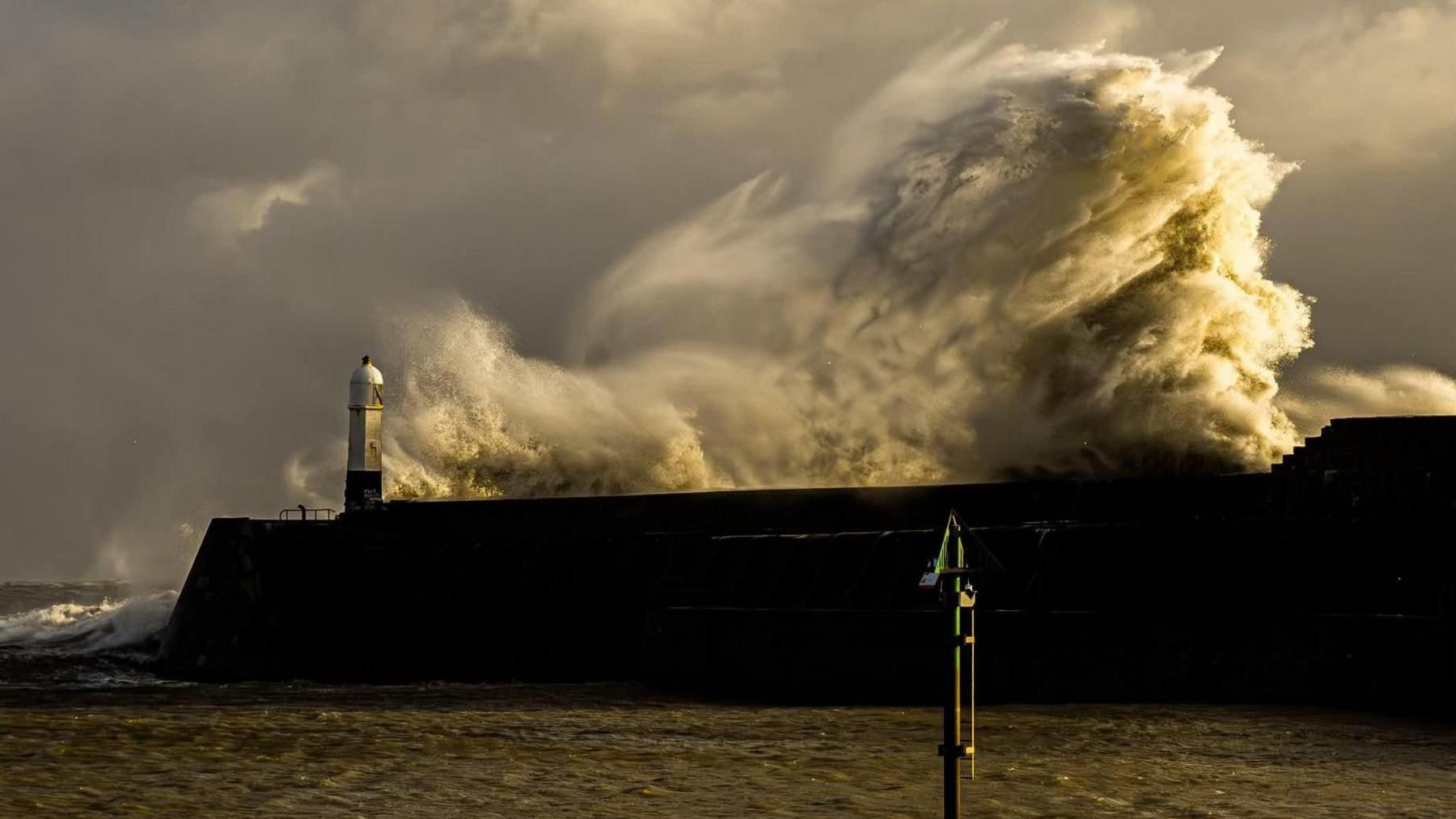 Waves crashing against a breakwater surrounded by sea, under a heavy grey sky 